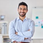Young happy mixed race businessman standing with his arms crossed working alone in an office at work. One expert proud hispanic male boss smiling while standing in an office